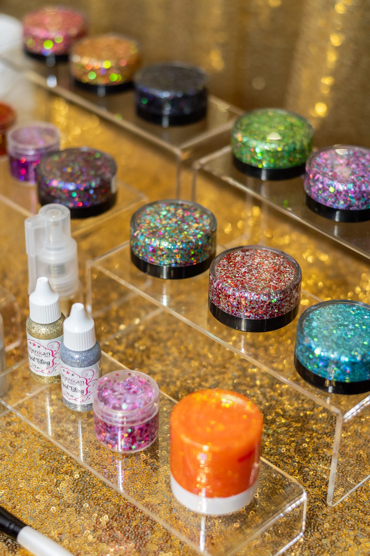 Jars of chunky multicolor glitter and small glitter bottles arranged on clear risers over a gold sequin backdrop.