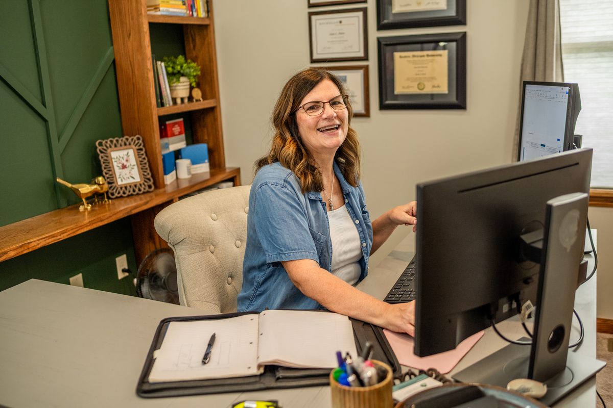 A smiling woman with glasses in a denim shirt works at a desk with a large monitor, a binder and a pen cup, with framed certificates and a green accent wall behind her.