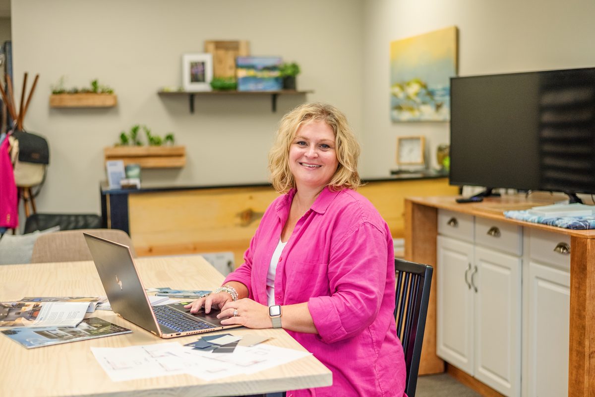 A smiling woman in a bright pink shirt types on a laptop at a light wood table with printed materials and swatches, in a bright office.