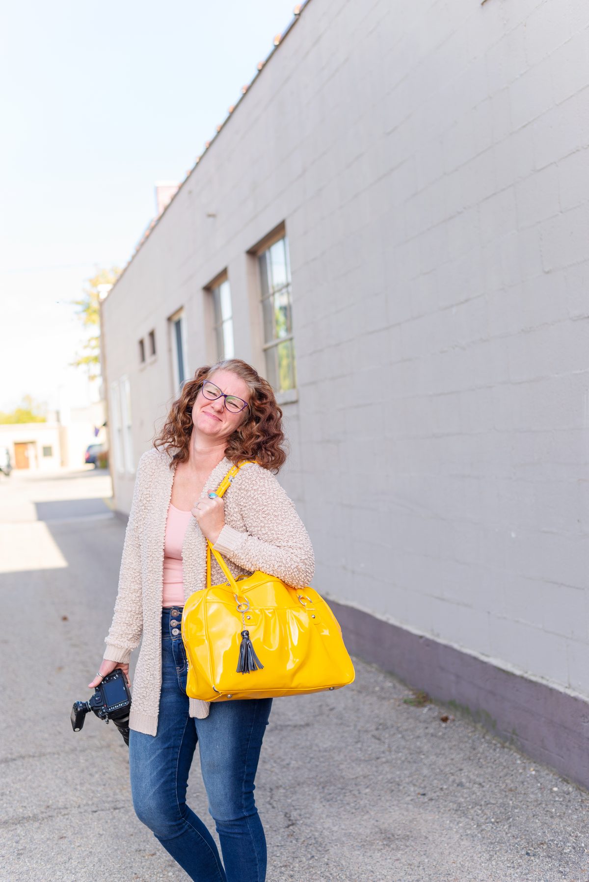 A woman with curly auburn hair and glasses in a cream cardigan walks along a pale brick alley with a yellow bag on her shoulder and a camera in her hand.