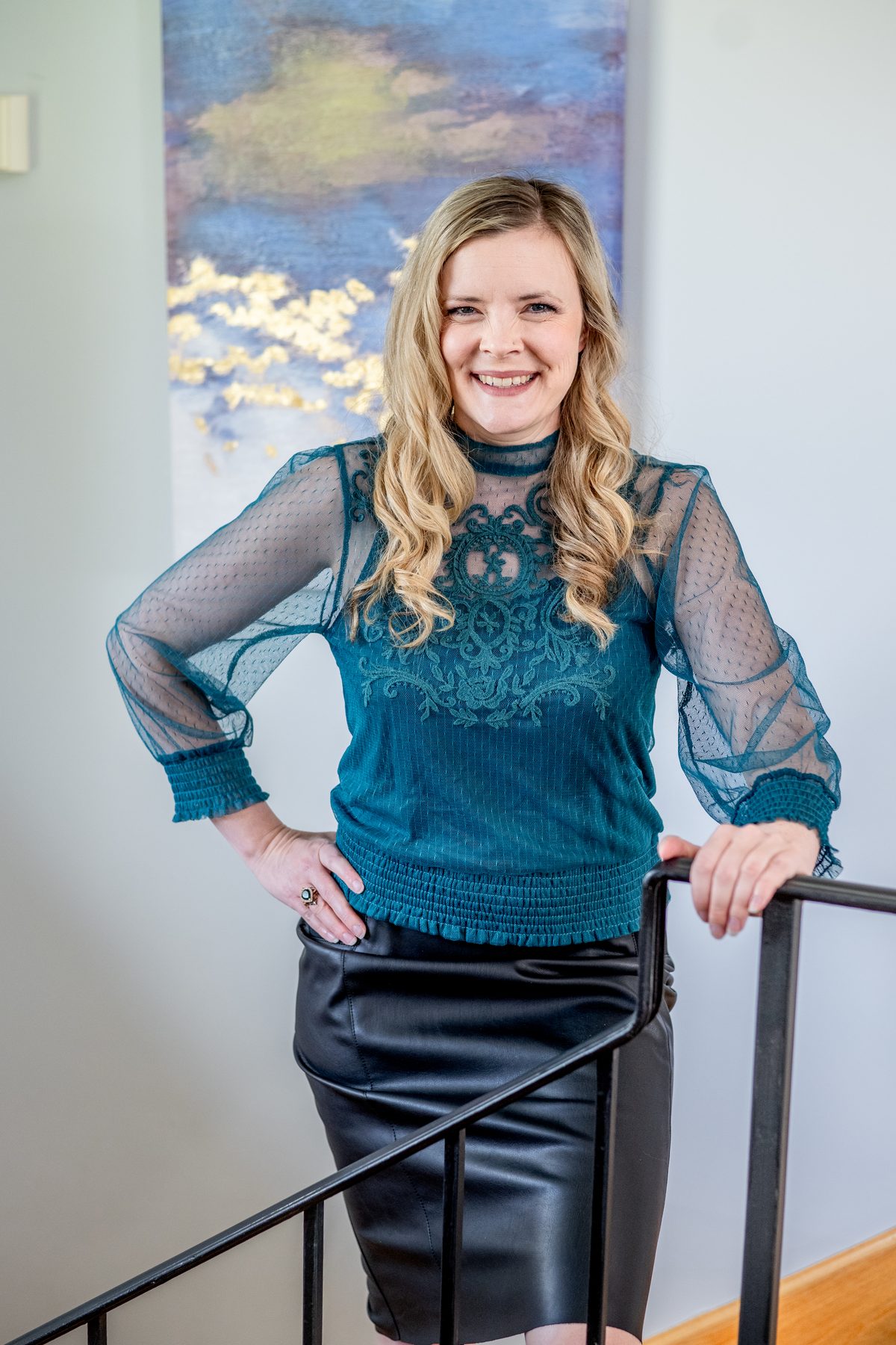 A woman with long blonde wavy hair in a teal lace blouse smiles with one hand on her hip and the other on a stair railing.