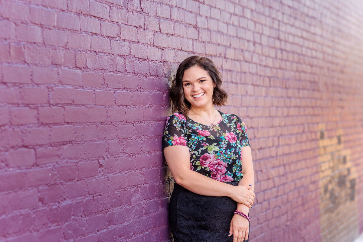 A smiling woman with short dark hair in a floral top leans against a pink and purple painted brick wall.