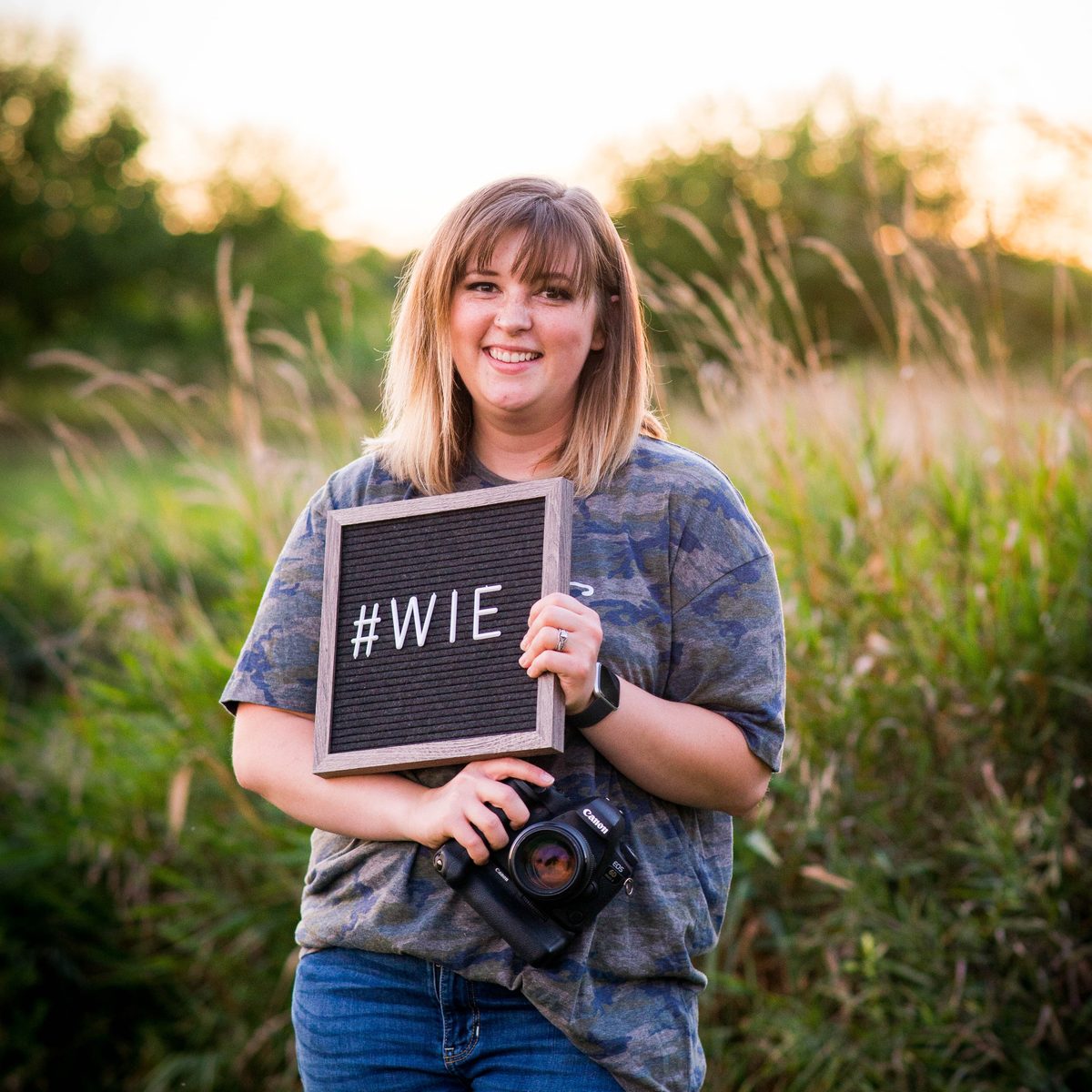 A smiling woman with bangs in a camouflage tee holds a letter board and a camera in a grassy field at golden hour.