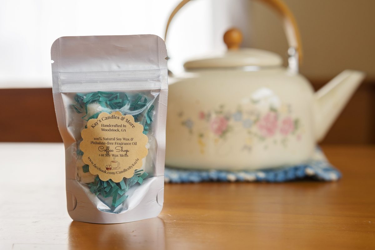 A clear-front silver pouch of soy wax melts with a gold label, standing on a wooden table in front of a cream floral teakettle.