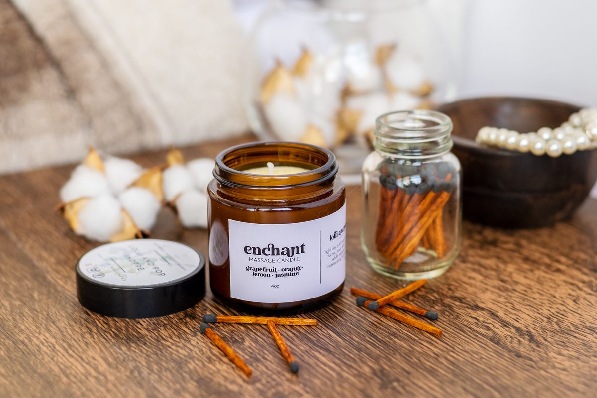 An amber jar candle with a cream label on a wooden table, beside a jar of matches, loose matches, cotton stems, a small tin and a bowl of pearls.