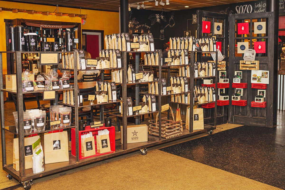 Wheeled metal shelves in a coffee shop stocked with bagged coffee, boxed gifts and mugs, in front of a chalkboard wall and a dark display door.