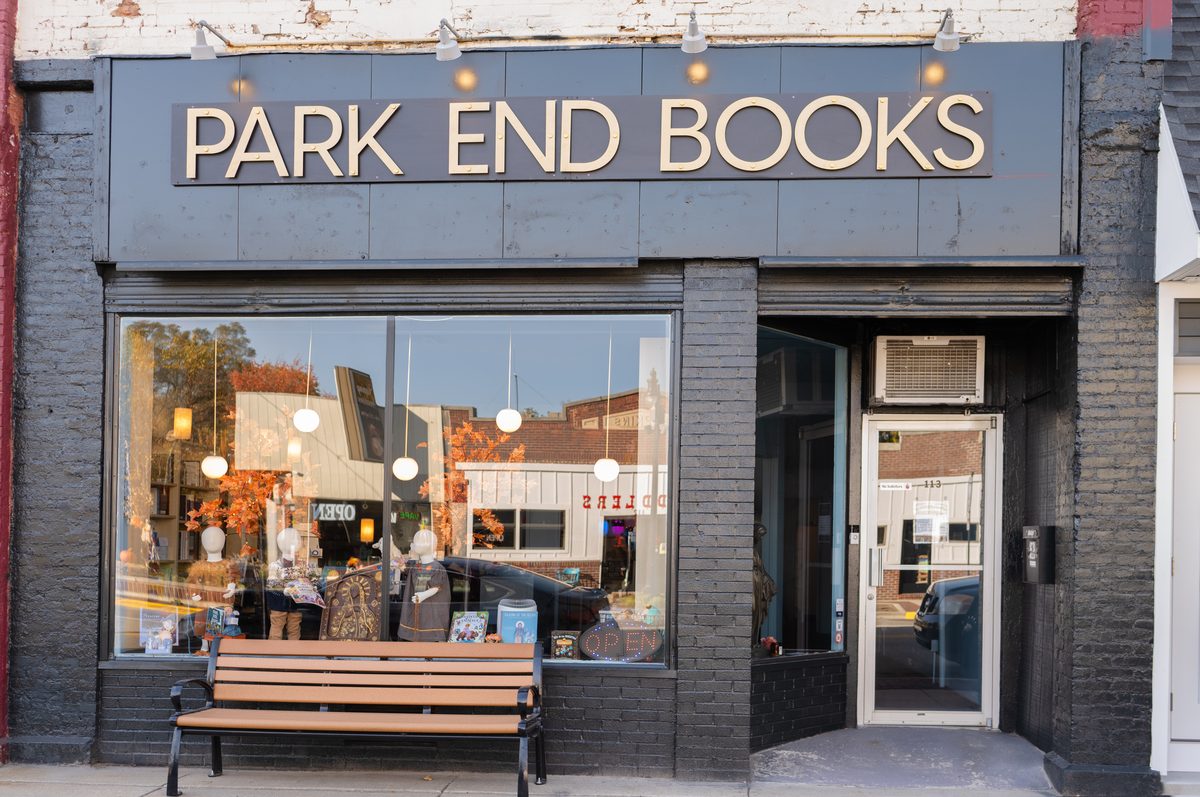The front of Park End Books: a dark brick storefront with a large window of seasonal displays, a wooden bench, an open sign and the entrance door.