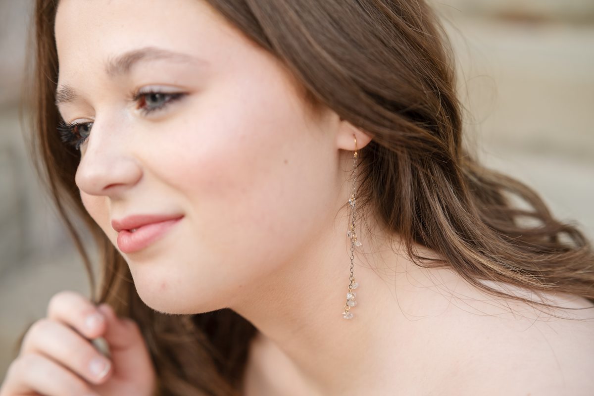 A woman touching her ear to show a gold earring with black beads and a moonstone drop, with a small leaf tattoo on her wrist.