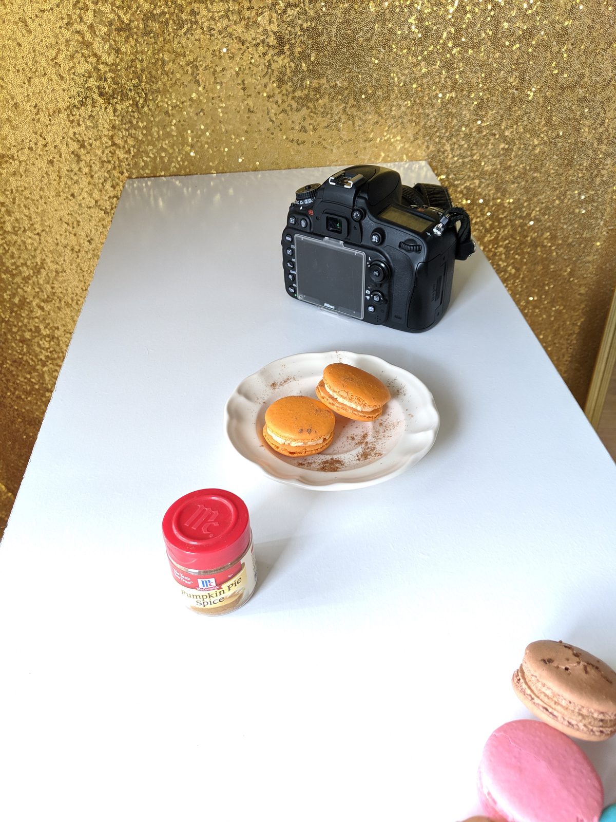 A camera, a plate of orange macarons and a jar of pumpkin pie spice on a white table in front of a gold glitter backdrop, a product shoot being set up.