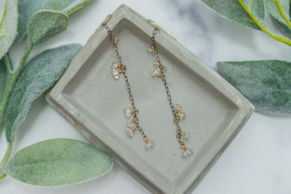 A pair of long dangling earrings with clear crystal clusters and gold accents, laid on a grey concrete tray among soft green lamb's-ear leaves.