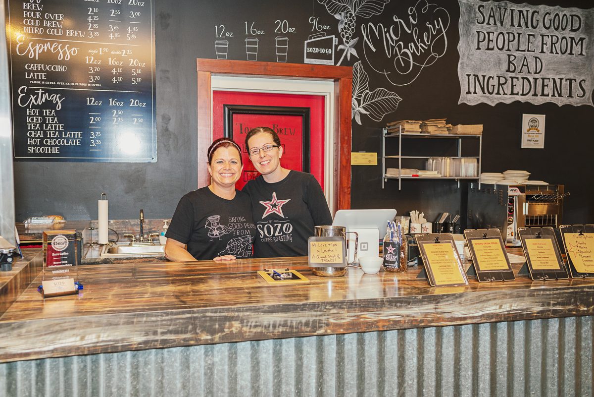 Two coffee shop employees in matching black shirts smiling together behind their counter, with the menu board and shelves behind them.