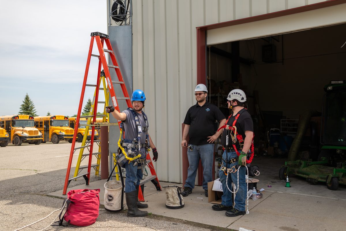 A worker in a blue helmet and safety harness stands beside an orange ladder while two colleagues talk by an open shop door, with school buses in the lot behind.