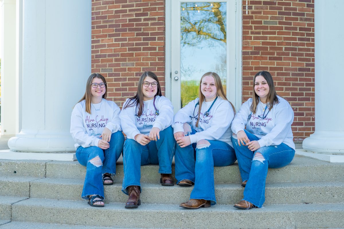 Four nursing students in matching sweatshirts and jeans sit side by side on the steps of a brick building with white columns.
