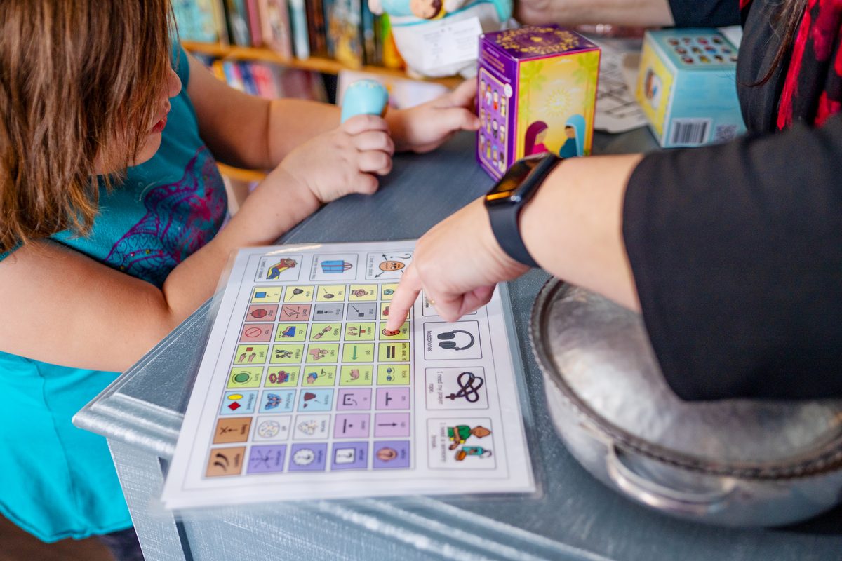 Close view of two people leaning over a gray table as one points to a printed communication board, with colorful boxes and bookshelves behind.