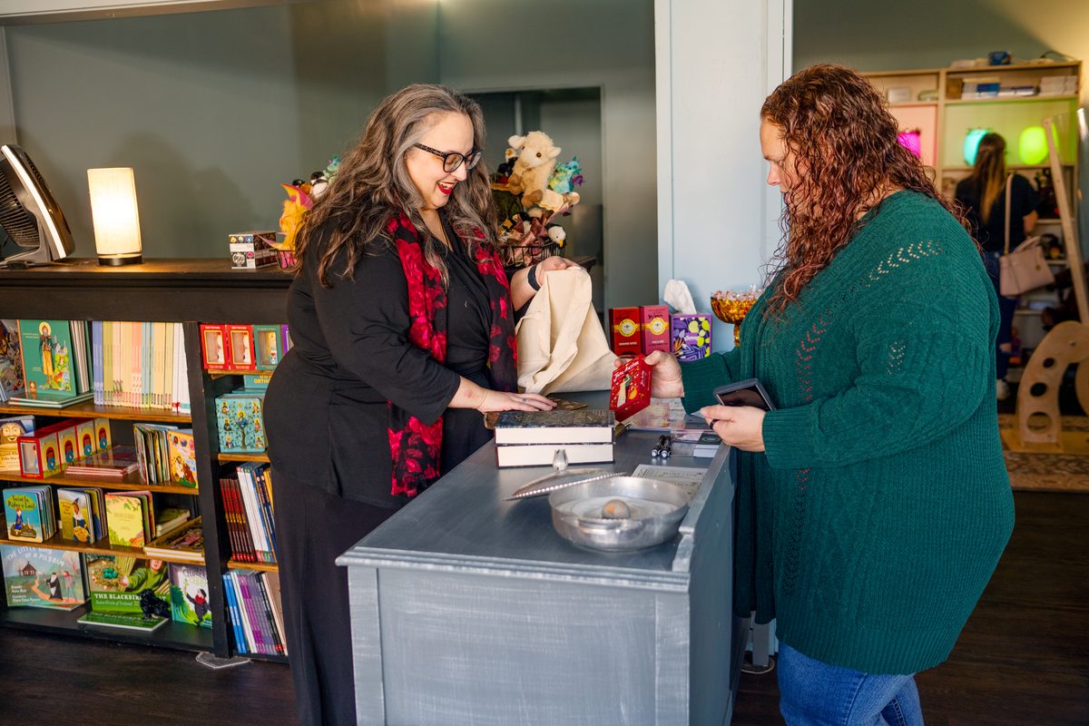 A shop owner in a black cardigan rings up a customer in a green sweater at a counter in a toy and book store.