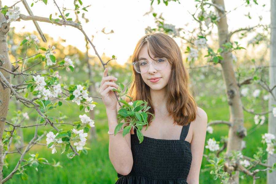 A young woman in a black dress standing amidst blooming trees, holding a branch with apple blossoms.