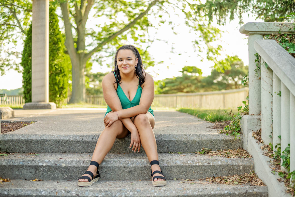 a high school senior girl in a green dress sits on outdoor stone steps, smiling in a leafy park setting with trees and a pathway in the background for senior photo shoot.