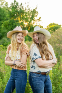 Two smiling women wearing cowboy hats and tie-dye shirts stand back-to-back with arms crossed, outdoors with greenery in the background.
