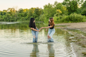 Two high school girls standing in shallow water at a lakeside, holding hands. They are wearing jeans and black shirts, surrounded by trees and greenery.
