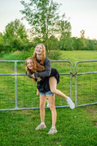 Two high school girls laughing, with one giving the other a piggyback ride in a grassy area near a metal fence. Trees and a clear sky are visible in the background.