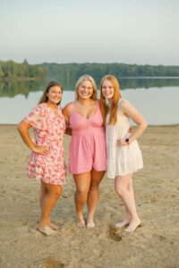 Three smiling high school seniors stand together on a beach, wearing summer dresses. A calm lake and trees are visible in the background. stand together on a beach, wearing summer dresses. A calm lake and trees are visible in the background.