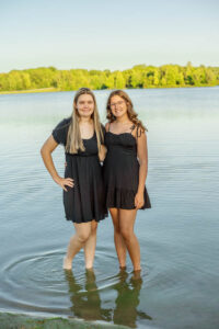Two high school seniors stand in shallow lake water, wearing black dresses, with trees in the background.