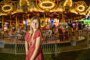 A high School Student in a red dress smiling in front of a lit-up carousel at night at the Ionia Free Fair.