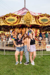 Three high school students stand in front of a colorful carousel at a fair. They are smiling and posing with their arms raised at night at the Ionia Free Fair.