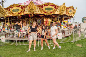Three teenage girls pose in front of a colorful, ornate carousel. Two stand with hands on hips, and they are smiling. The carousel features painted horses and bright lights at night at the Ionia Free Fair.