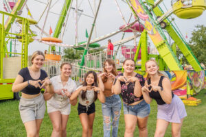 Six people stand in front of a colorful carnival ride, smiling and making heart shapes with their hands at night at the Ionia Free Fair.