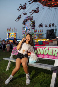 Girl sitting on a bench at a carnival, eating a dessert, with a swing ride and Ferris wheel in the background at night at the Ionia Free Fair.