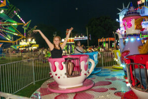 A person with raised arms sits in a spinning teacup ride at a brightly lit amusement park at night at night at the Ionia Free Fair.
