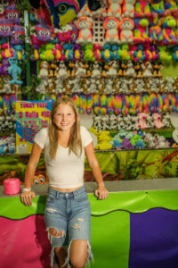 Person in a white shirt and denim shorts stands in front of a carnival game booth with colorful stuffed toys on display at night at the Ionia Free Fair.