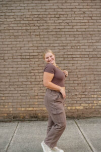 A high school senior in a brown shirt and cargo pants poses smiling in front of a brick wall, with one hand on their hip and the other touching their collar for a Monochromatic photo shoot.