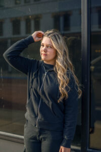 A high school senior with long hair wearing a dark hoodie stands outside a building, touching their hair and gazing into the distance. The window behind reflects some light for a Monochromatic photo shoot.