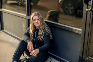 A high school senior with long hair sits on the sidewalk leaning against a glass storefront. She is wearing a black outfit and looking directly at the camera for a Monochromatic photo shoot.