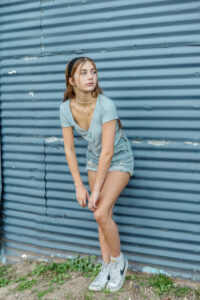 A high school student in a light blue shirt and denim shorts leans against a corrugated metal wall, looking to the side, with sneakers on grassy ground for a Monochromatic photo shoot..