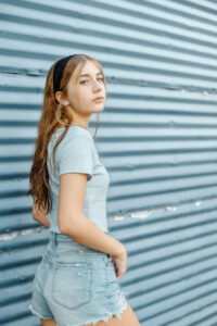 A high school student with long hair and headband wearing a light blue shirt and denim shorts stands in front of a corrugated metal wall, looking towards the camera for a Monochromatic photo shoot..
