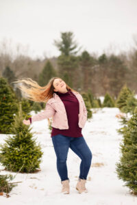 A high school student standing in a snowy field with evergreen trees, wearing a pink jacket and jeans, hair flowing to the side.
