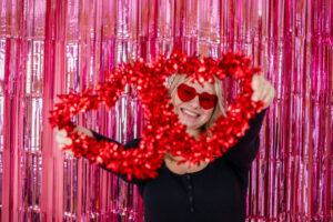 Female High School Senior holding a red heart decoration, wearing red heart-shaped sunglasses, smiling, in front of a pink tinsel backdrop for a Valentine's Day photo shoot.
