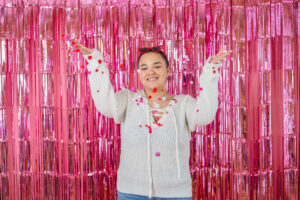 Female High School student in a white sweater stands in front of a shiny pink backdrop, smiling and holding red pom-poms, with sunglasses perched on their head for a Valentine's Day photo shoot.