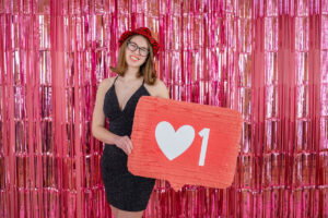 Smiling Female High School Senior in a black dress and red hat holds a sign with a heart and the number 1, standing before a pink tinsel backdrop for a Valentine's Day photo shoot.