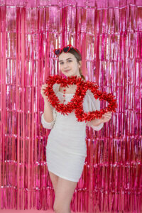 Female High School student in a white dress holding red heart-shaped decorations in front of a pink metallic fringe backdrop for a Valentine's Day photo shoot.