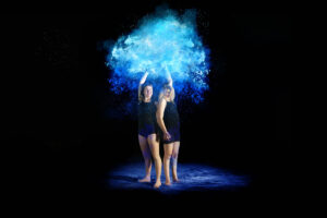Two people in black attire stand with raised arms as vibrant blue flour powder bursts above them against a dark background.