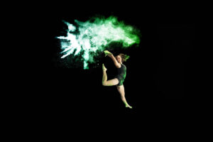 Student dancer in mid-air pose on a black background with green flour powder exploding behind them.