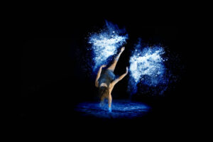 A student performs a handstand on one hand with blue and white flour surrounding them against a dark background.