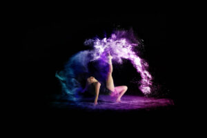 A person performs a dynamic dance pose, surrounded by swirling clouds of purple and blue powder against a black background.