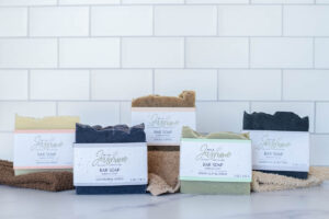 Five bar soaps with labeled packages are displayed on a counter with a white tiled backsplash. Each soap bar is a different color, and they are placed on cloths.
