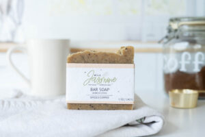Bar of spiced coffee soap labeled "Wild Jasmine" on a cloth, with a jar of coffee grounds and a white mug in the background.